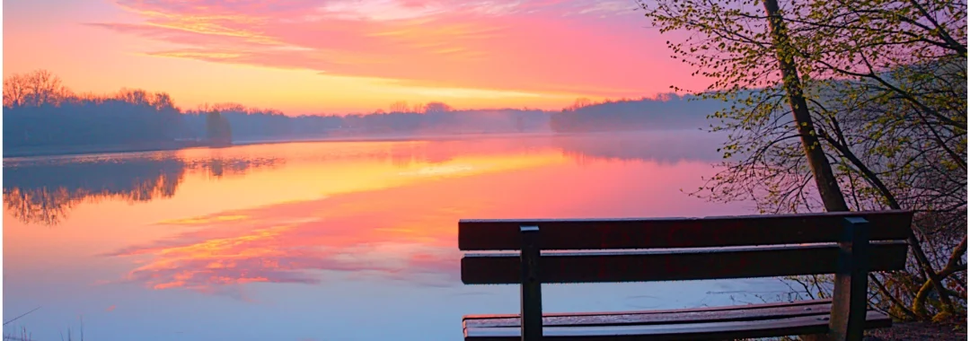 bankje aan een meer. De zon wolken aan de horizon worden prachtig aangelicht door de nog op te komen zon.