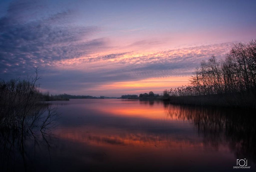 Een sereen meer bij zonsondergang, met een feloranje en paarse lucht die weerspiegelt in het kalme water, omlijst door de silhouetten van bomen en riet aan de oevers.