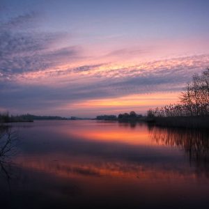 Een sereen meer bij zonsondergang, met een feloranje en paarse lucht die weerspiegelt in het kalme water, omlijst door de silhouetten van bomen en riet aan de oevers.