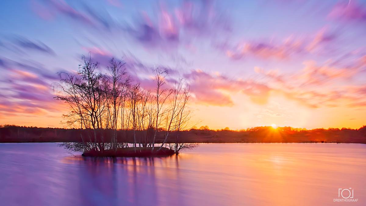 Zonsondergang boven een kalm meer, met silhouetten van bomen die uit het water oprijzen, oranjeroze gloeiende wolken en fel paarse weerspiegelingen; een serene sfeer.