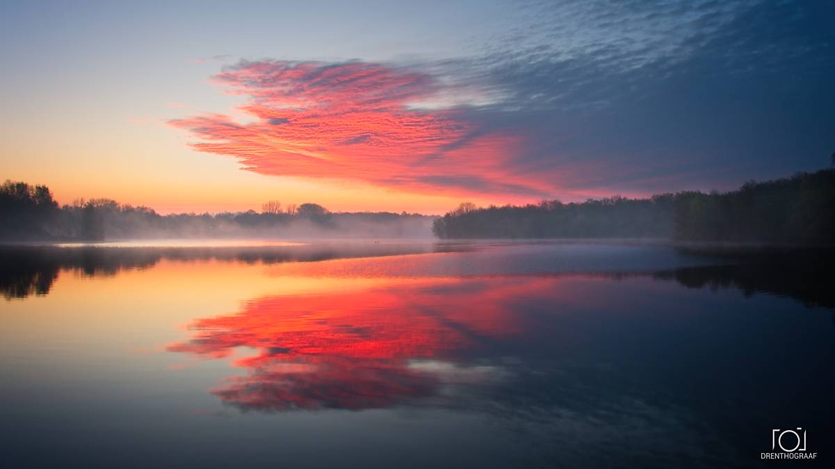 Een adembenemende zonsopgang boven een mistig meer met levendige rode wolken die in het water weerspiegelen, vastgelegd in een serene landschapsfoto van Drenthograaf.