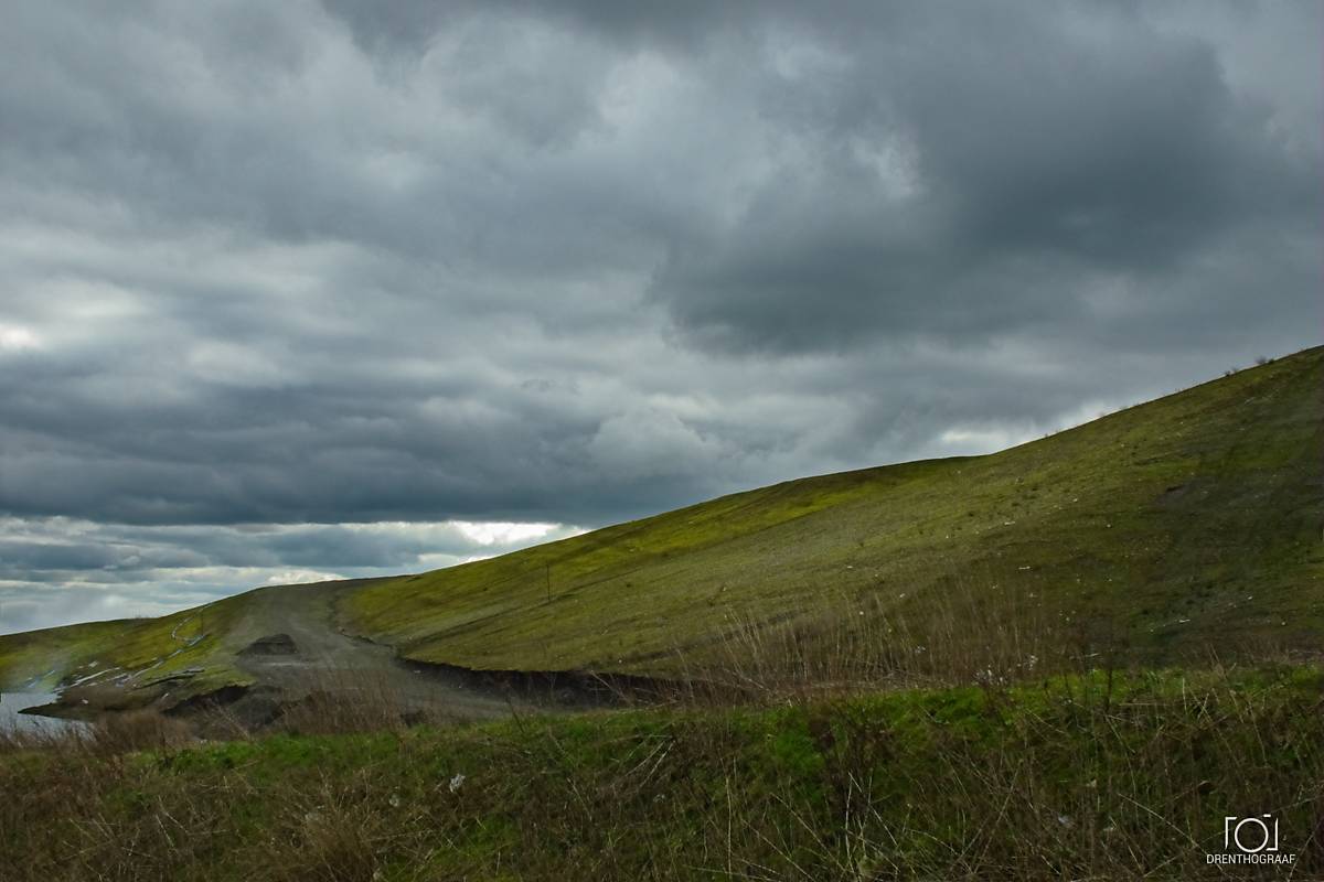 Een met gras begroeide heuvel loopt omhoog onder een dramatische, zware, donkergrijze bewolkte hemel, met een zandpad dat langs de voet slingert, vlakbij een klein wateroppervlak.